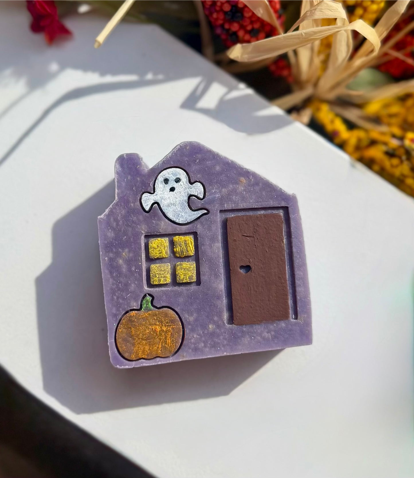A purple Halloween-themed house-shaped soap with a white ghost, yellow window, brown door, and orange pumpkin stamped on the front of the soap, photographed on a white surface with warm natural light and blurred dried flowers in the background.