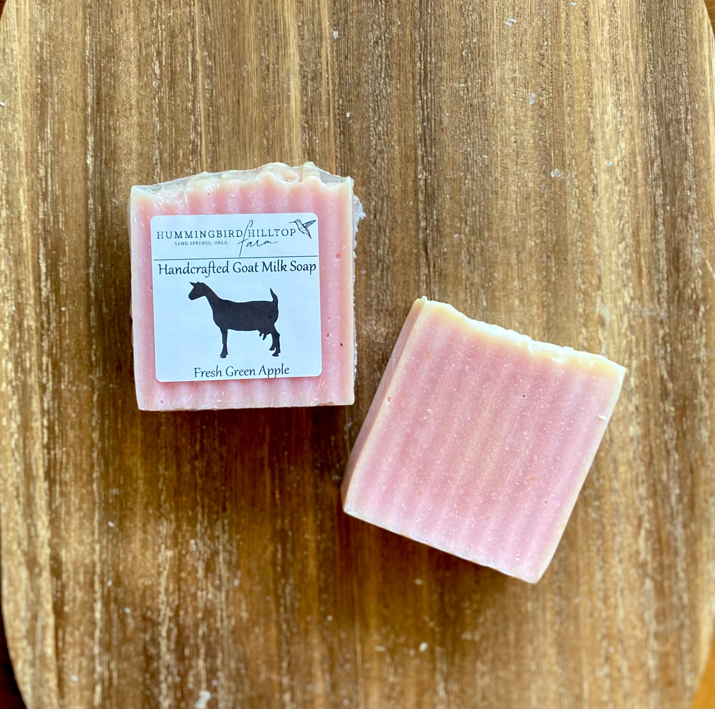 Two light pink bars of soap on a wooden background. One bar is shrinkwrapped and labeled.