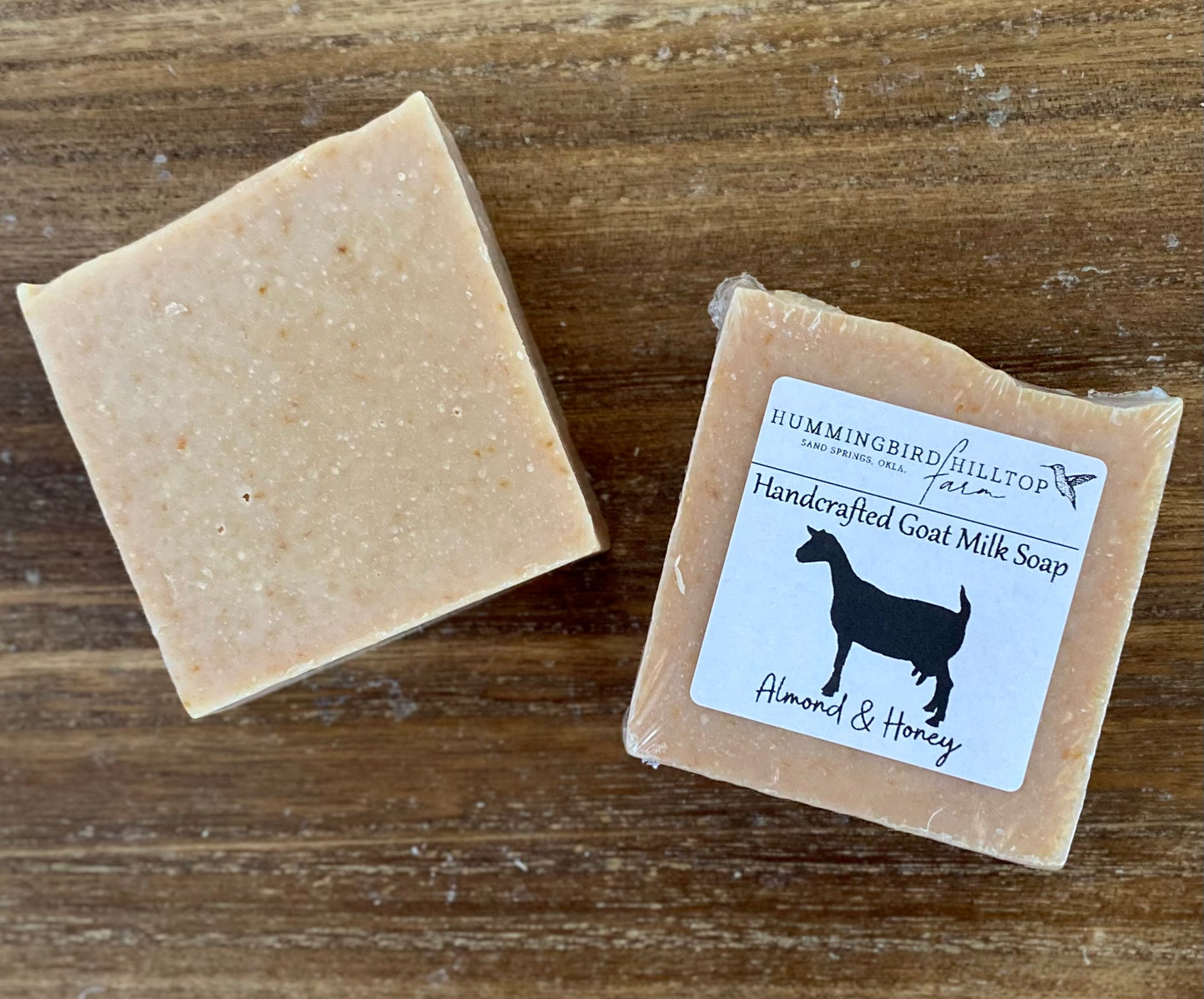 Two light brown bars of Almond and Honey Goat Milk Soap on a wooden surface. One bar is unwrapped and the other bar is shrinkwrapped and labeled.