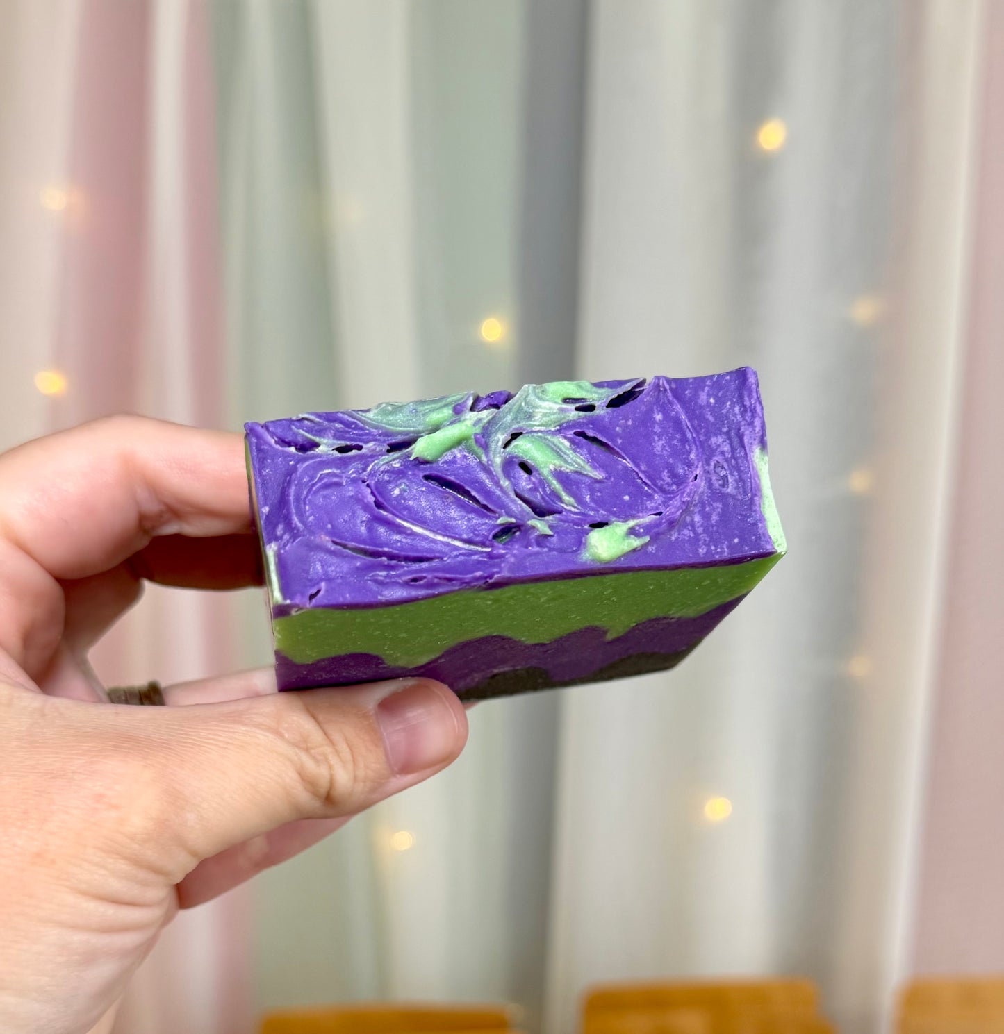 A hand holding a rectangular handmade soap bar with a swirled purple and green top.