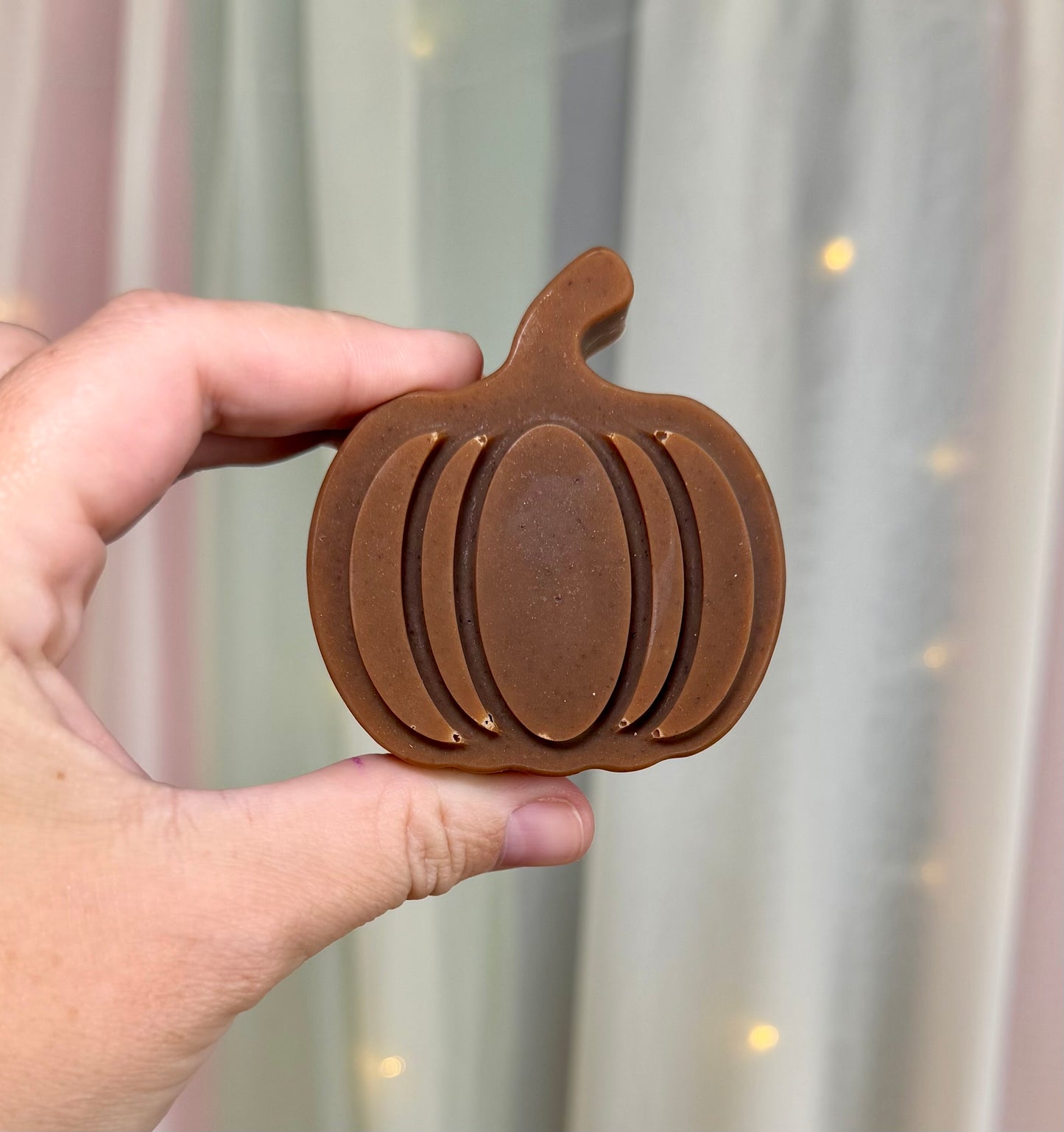 A hand holding a pumpkin-shaped brown soap bar against a soft-focus background of curtains and warm string lights.