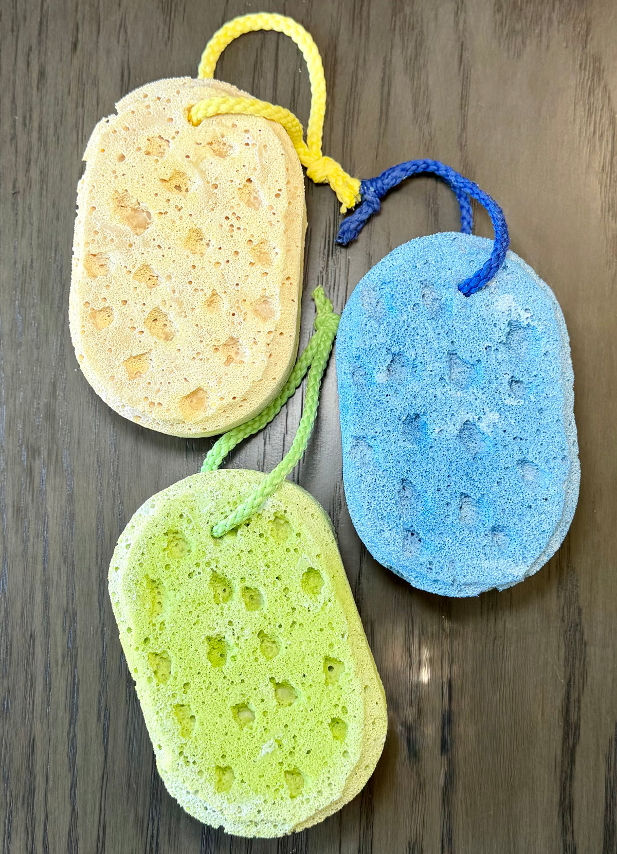 3 sponges in yellow, blue and green colors with hanging cords on a wooden background.