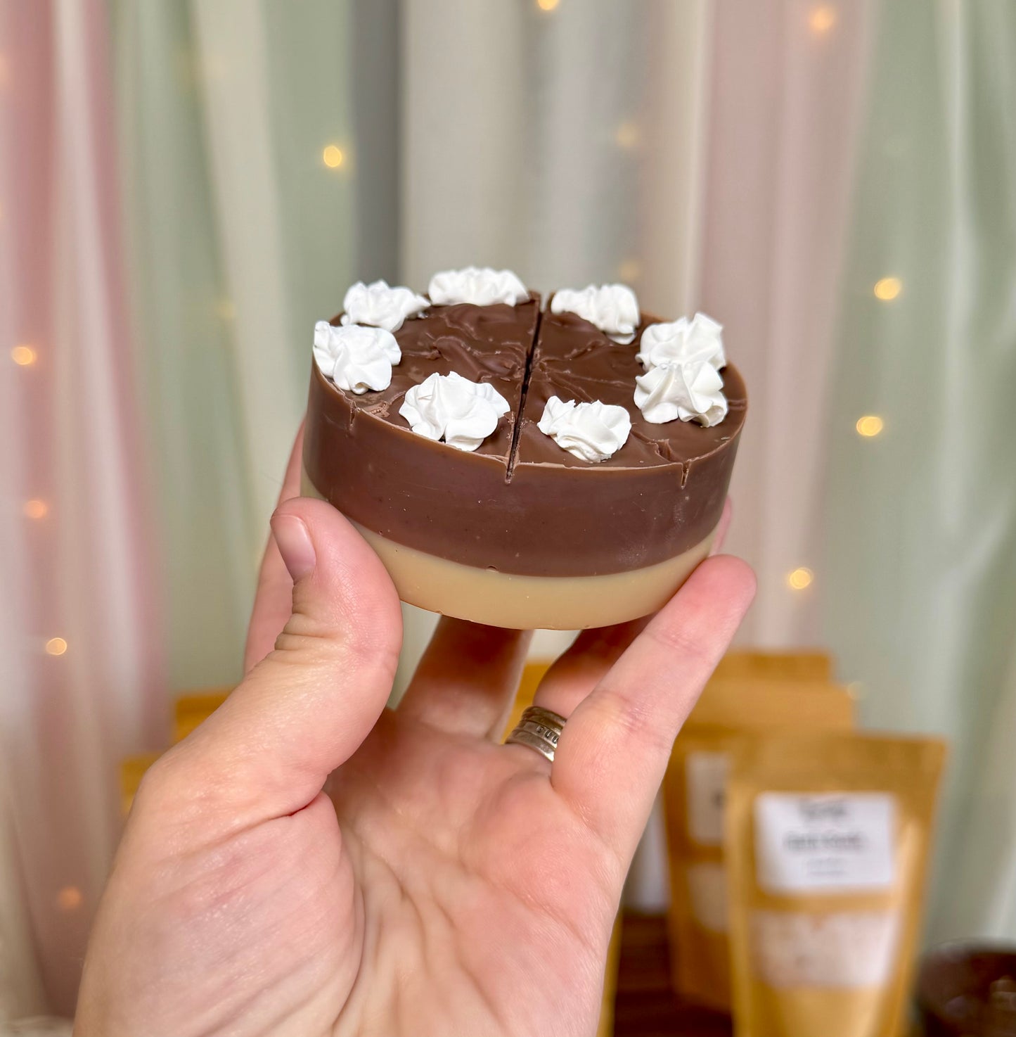 A hand holds a small round soap shaped like a pie, with a dark brown top layer, a beige bottom layer, and white whipped cream dollops on top. The background shows soft draped curtains and warm string lights.