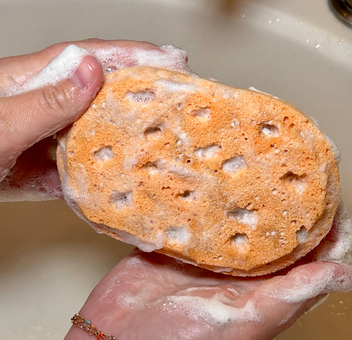 Two hands holding orange colored sponge with lather and bubbles on sponge and hands.