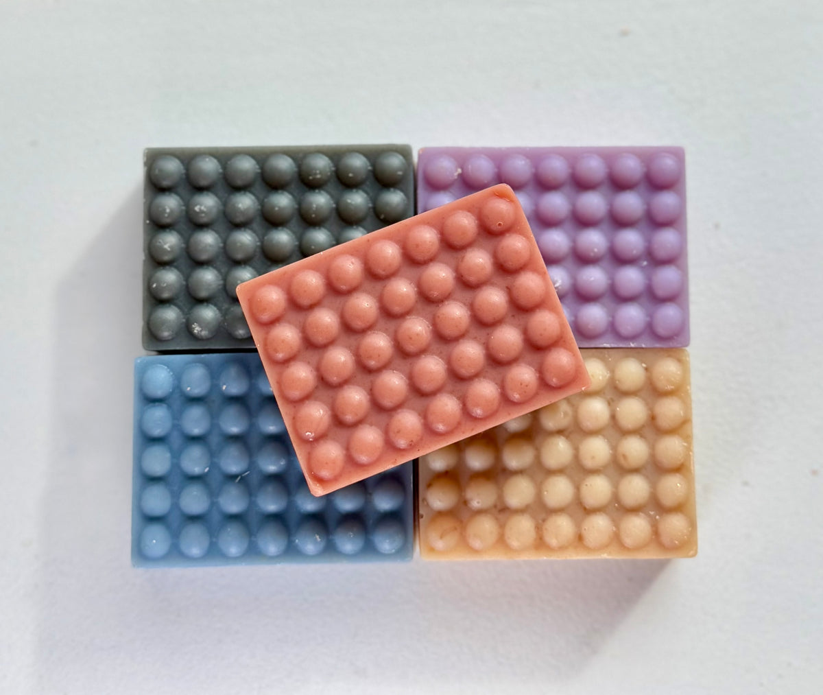 5 mini bars of soap in 5 different colors (grey, purple, blue, pink, and brown) stacked on a white background