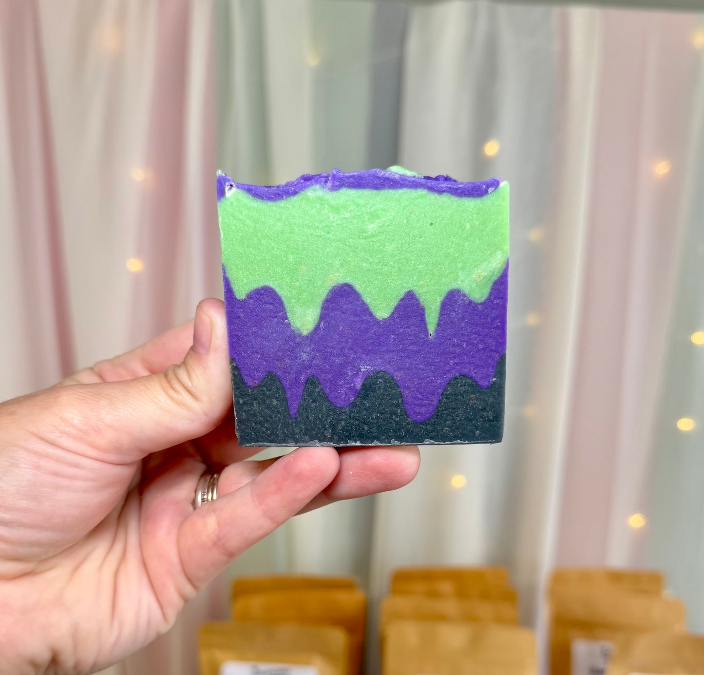 A hand holds a square layered soap bar with green, purple, and black drip designs. The background shows soft pink and white curtains with warm string lights.