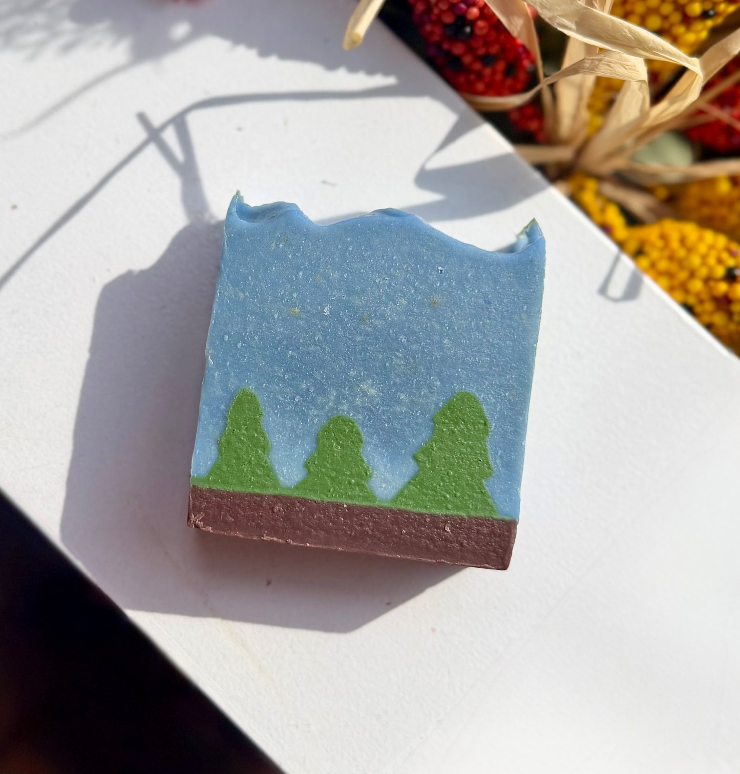 A rectangular handmade soap bar with a landscape design, featuring a light blue sky, green pine trees, and a brown ground layer, displayed on a white surface with natural shadows.