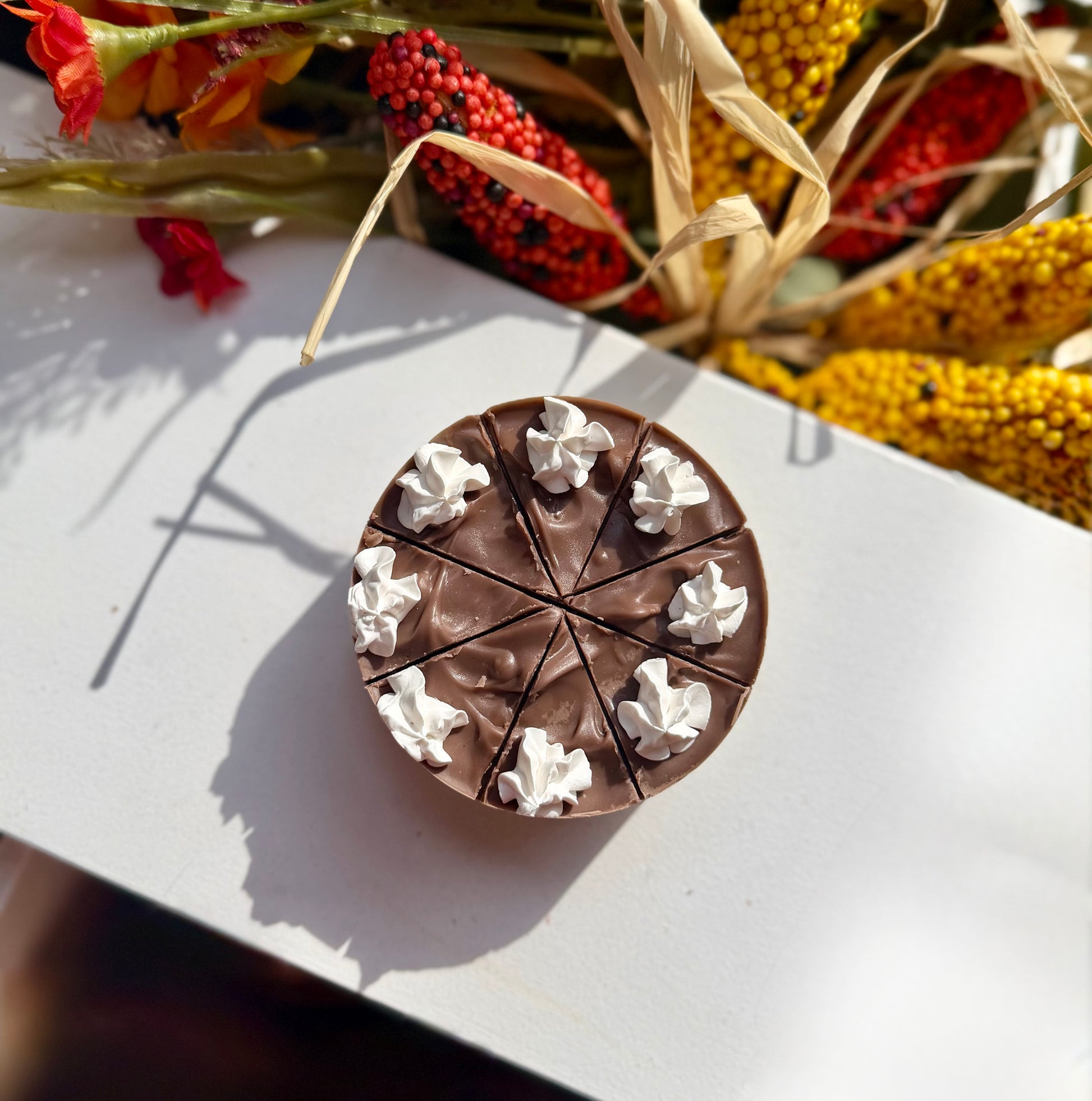 A round soap that looks like pie topped with white whipped cream dollops, photographed on a white surface with dried corn and autumn flowers in the background.