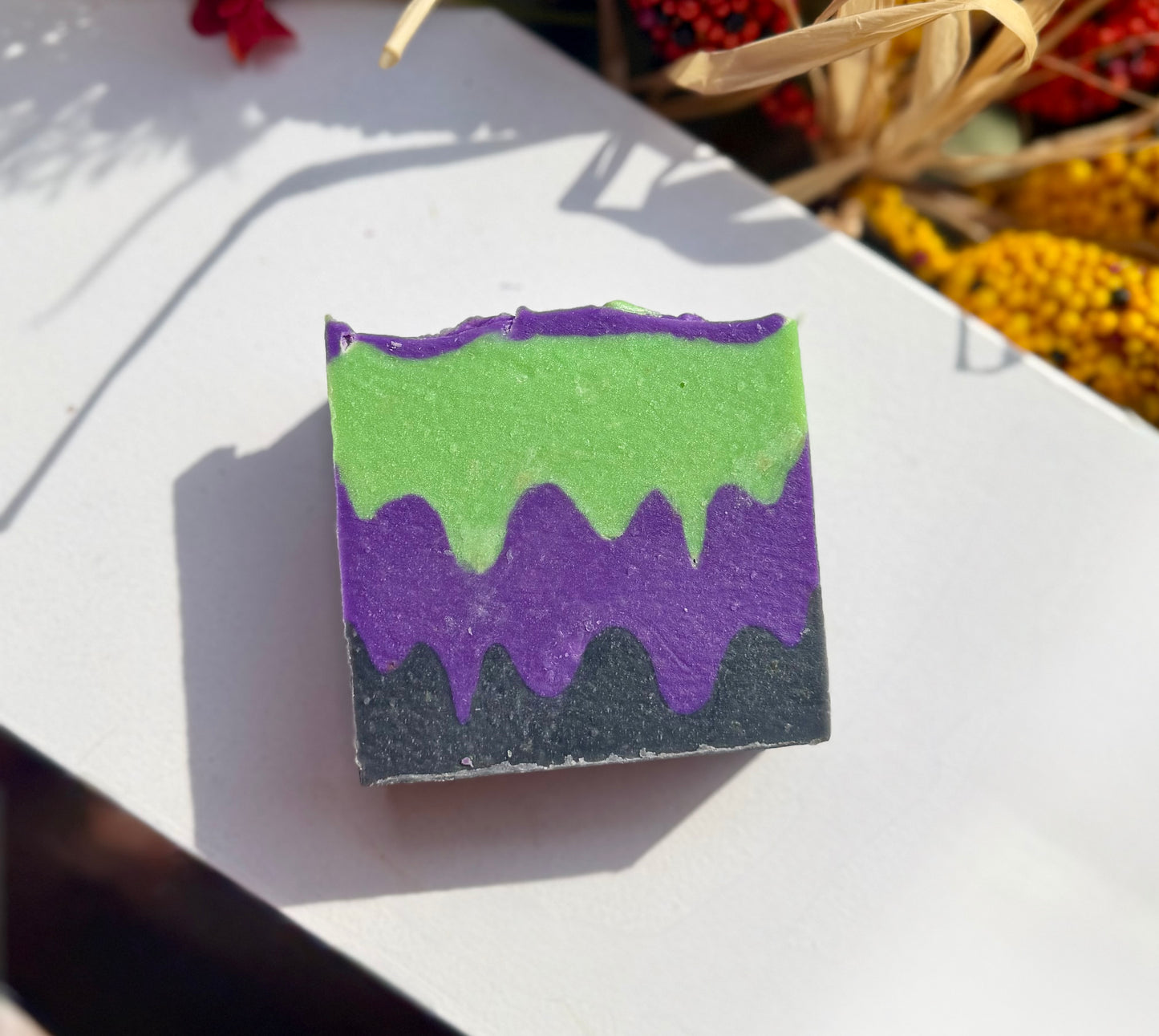 A rectangular bar of handmade soap with a three-color drip design, featuring lime green on top, purple in the middle, and a dark charcoal gray base, resting on a white surface with decorative dried flowers in the background.