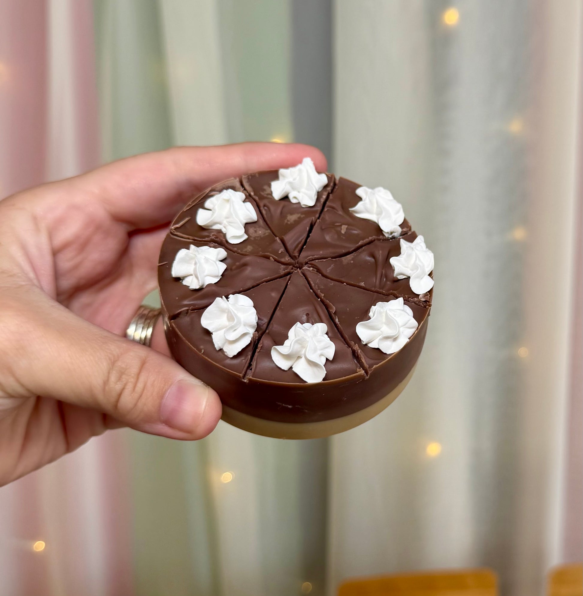A hand holds a small round soap shaped like a pie, with a dark brown top layer, a beige bottom layer, and white whipped cream dollops on top. The background shows soft draped curtains and warm string lights, with blurred pouches behind.