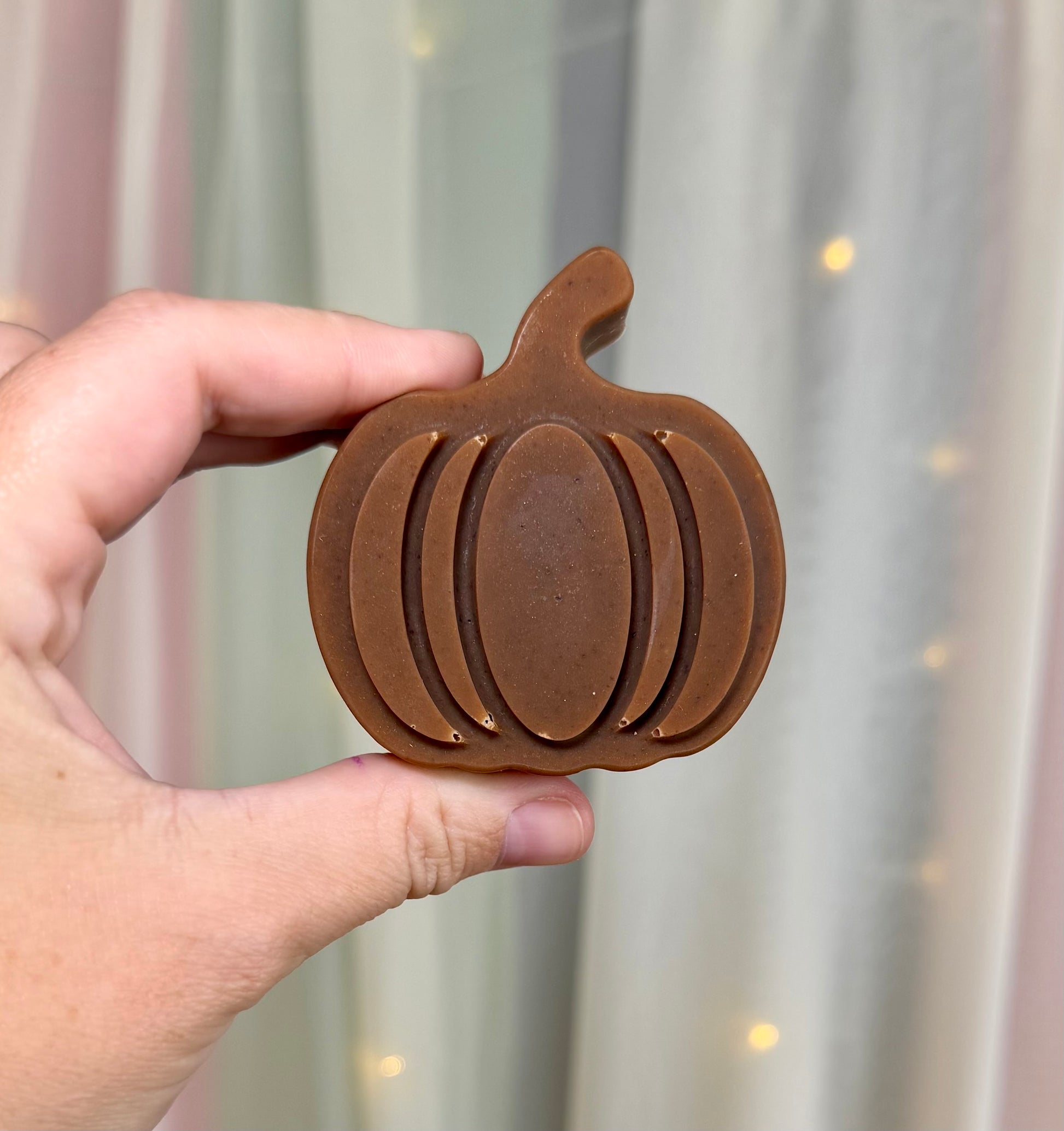 A hand holding a pumpkin-shaped brown soap bar against a soft-focus background of curtains and warm string lights.