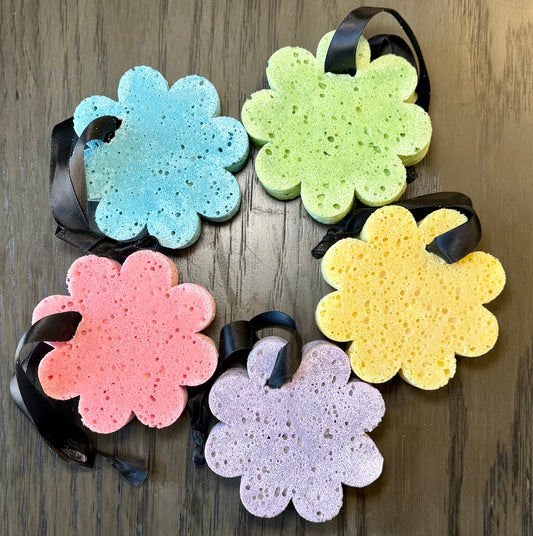 5 flower shaped sponges colored in blue, green, pink, yellow, and purple with black hanging cords on a wooden background.